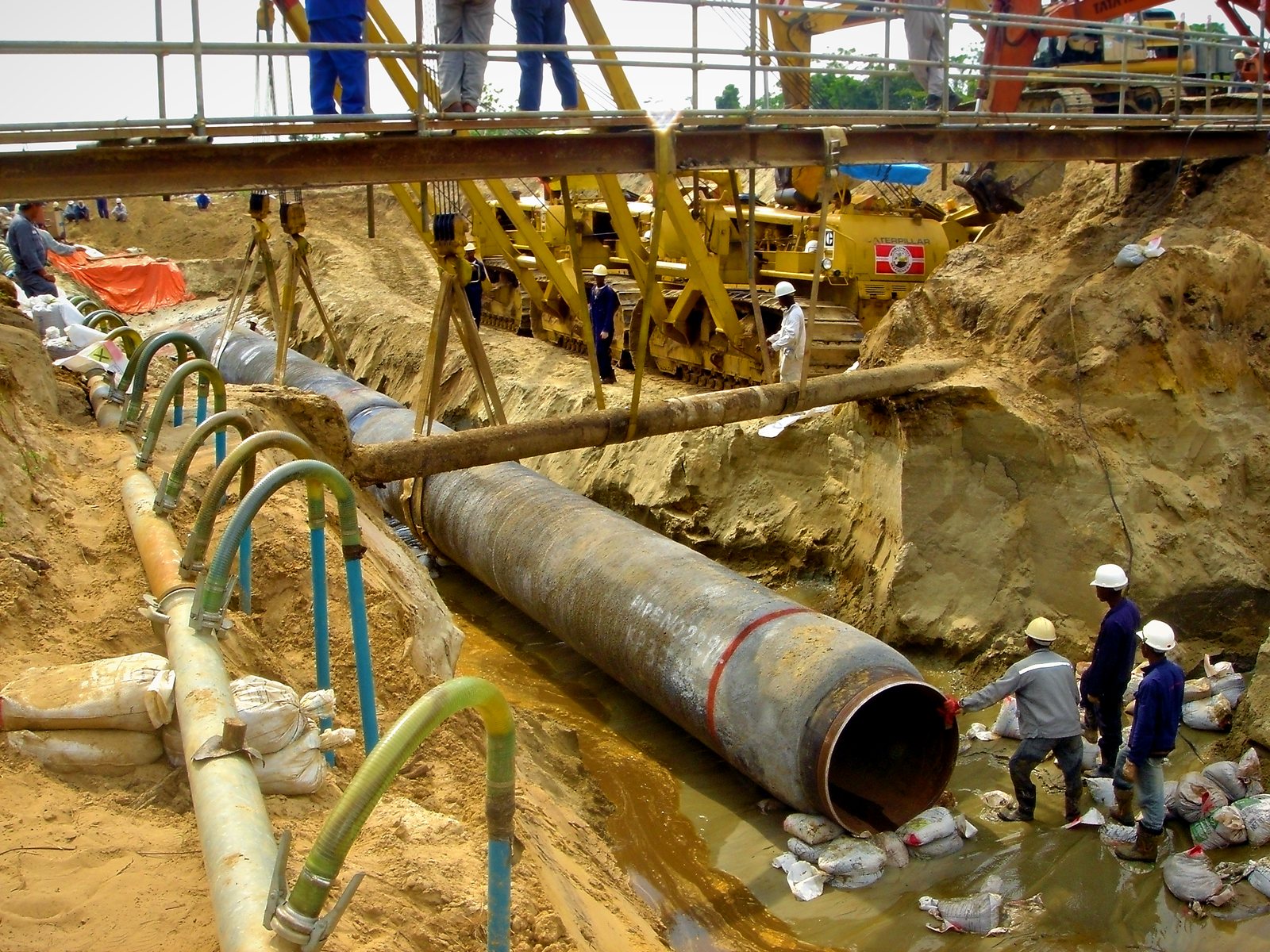Pipeline crossing: pipe in a dewatered trench beneath a temporary access bridge, with sideboom support